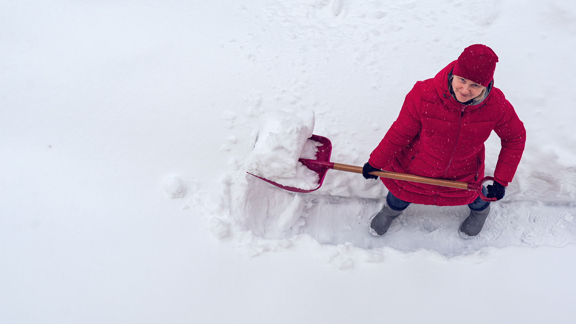 Nainen tekemässä lumitöitä talvella lumilapion kanssa punainen takki ja pipo päällä